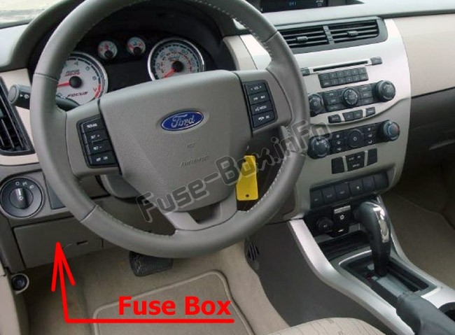 Passenger compartment fuse box location in a 2010 Ford Focus, situated below the steering wheel and to the left of the brake pedal, after removing the under-steering wheel cover.